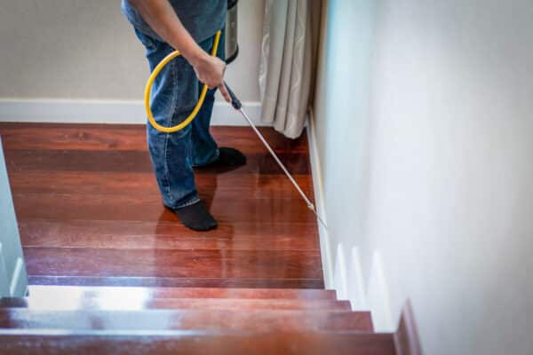 A person in jeans and black socks uses a sprayer with a yellow hose at the base of a wall by wooden stairs, likely for pest control.